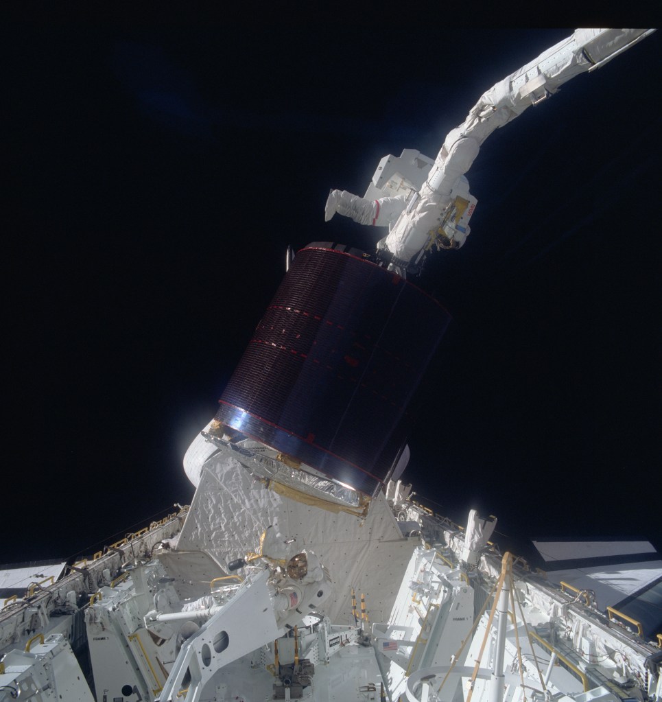 Astronaut Joseph P. Allen IV, top, hangs onto a stinger device as Astronaut Dale A. Gardner in the cargo bay of Discovery waits to assist in the berthing of the previously stranded satellite. The end effector of the remote manipulator system (RMS), controlled from inside the Discovery's cabin, grasps a special grapple point to Allen's right.