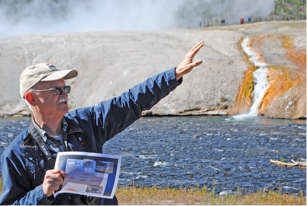 Des Marais with a tan cap on, sunglasses, with a blue jacket on pointing with his arm to a geyser in Yellowstone National Park.