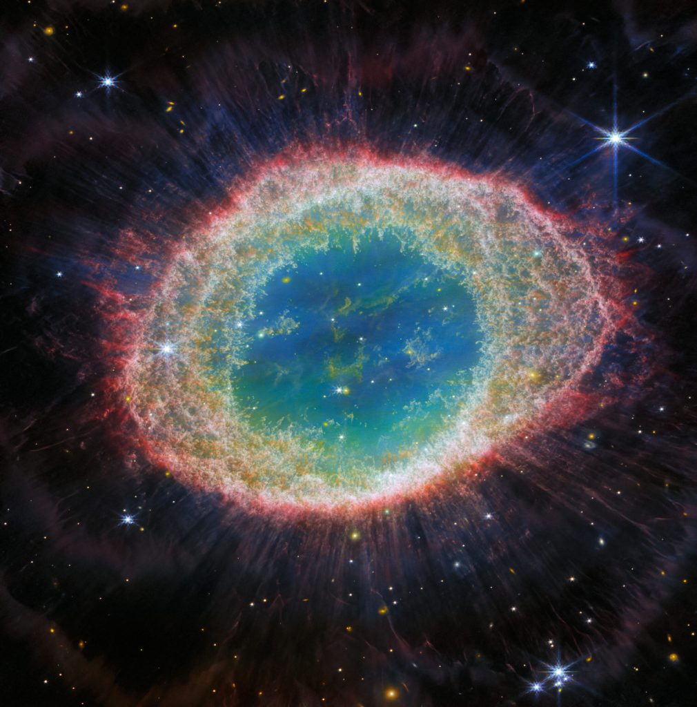 Alt text: This image of the Ring Nebula appears as a distorted doughnut. The nebula’s inner cavity hosts shades of blue and green, while the detailed ring transitions through shades of orange in the inner regions and pink in the outer region. The ring’s inner region has distinct filament elements.