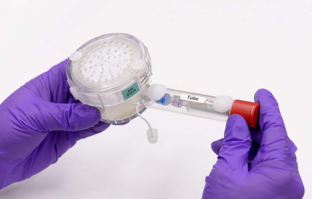 Purple-gloved hands seen holding experiment hardware with a round container at left, and plastic tube extending to the right.