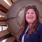 Adabelle Narvaez-Bostwick poses inside NASA Glenn’s Icing Research Tunnel. She is smiling, wearing a blue suit, and standing in front of a large, tan fan.