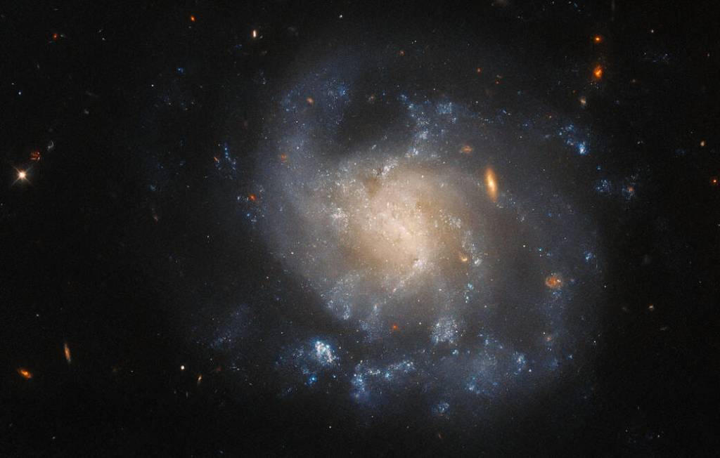 A irregularly-shaped spiral galaxy with difficult to distinguish arms. Its edges are faint, and the core has a pale-yellow glow. It is dotted with small, wispy, blue regions where stars are forming. A few stars and small galaxies are visible around it.