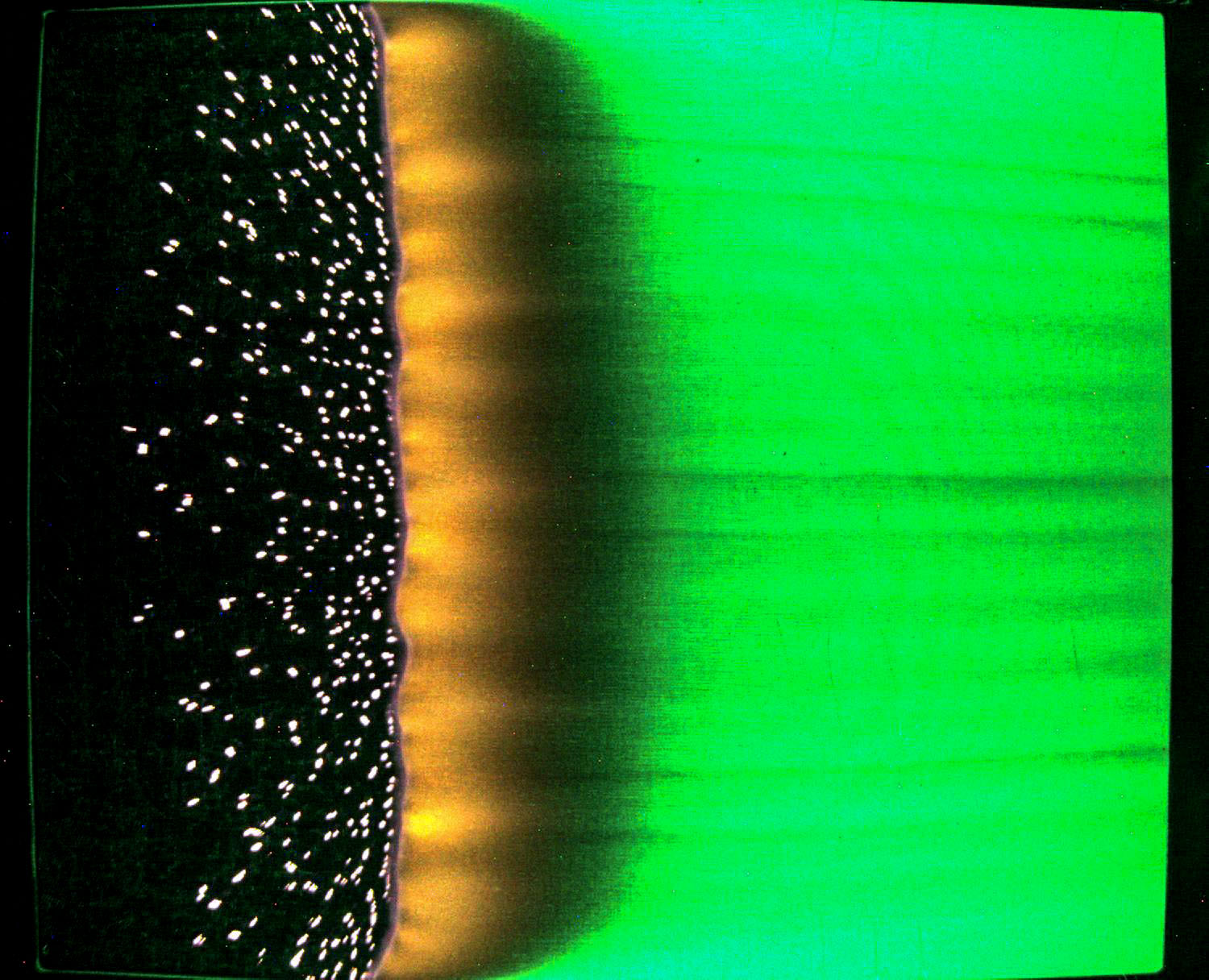 From left to right, this image has a band of black from top to bottom, a scattering of bright white specks of smoldering cotton like snowflakes, a band of orange flame, a small region of black where the cotton is beginning to char, and a wide band of unburned composite material that appears green because the sample is illuminated with green LED lights.