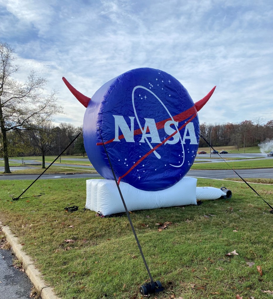 A photo of a large, inflatable NASA logo, which looks like a big blue circle with a red V-shape running diagonally across it, behind big white letters saying NASA and surrounded by stars. The word NASA and the red V are circled by a white oval representing a comet's path. The inflatable has the logo on both sides, so the point of the V on the back side is visible extending out the left side of the circle. The inflatable is staked to the ground outdoors with cables, and it is a slightly cloudy day with green-brown grass and trees in the background.
