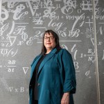 Maricela Lizcano stands inside NASA Glenn Research Center’s Aerospace Communications Facility. She is wearing a teal jacket and behind her there are mathematical equations on the wall.