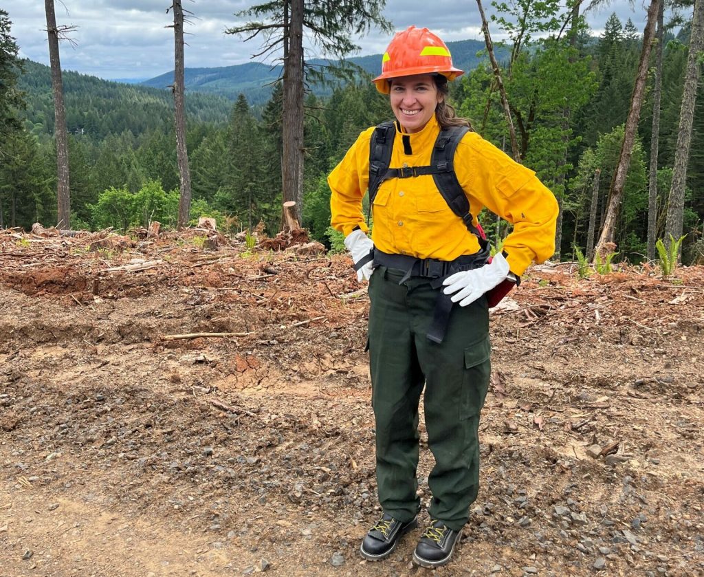 Former NASA intern Jenessa Stemke stands wearing a yellow long sleeve and dark green pants with protective gloves and helmet. She is smiling with her hands on her hips. Trees and mountains span the skyline. Credit: Paige Byassee