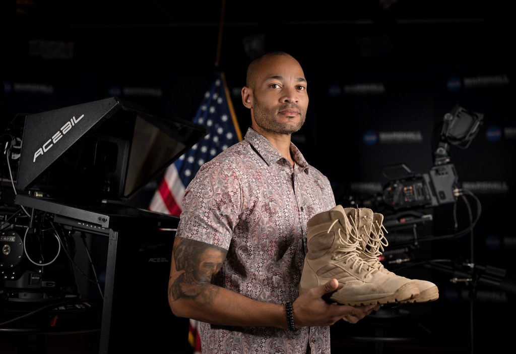 A man stands holding his combat boots in front of an American flag.