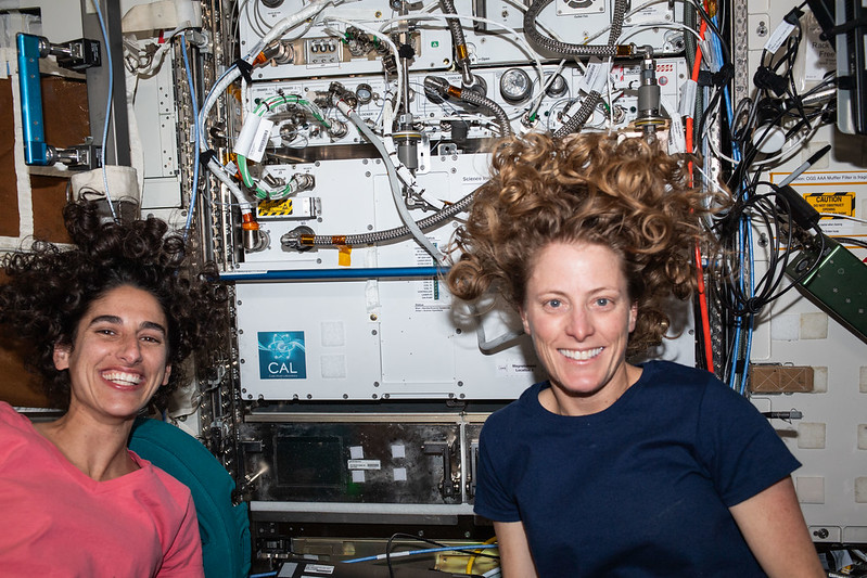 Moghbeli, left, wears a coral-colored t-shirt and O'Hara wears a dark blue t-shirt. Both are smiling at the camera as their curly hair floats around their heads. The white front of the Cold Atom Lab hardware, visible behind them, has multiple hoses and tubes attached to its front.