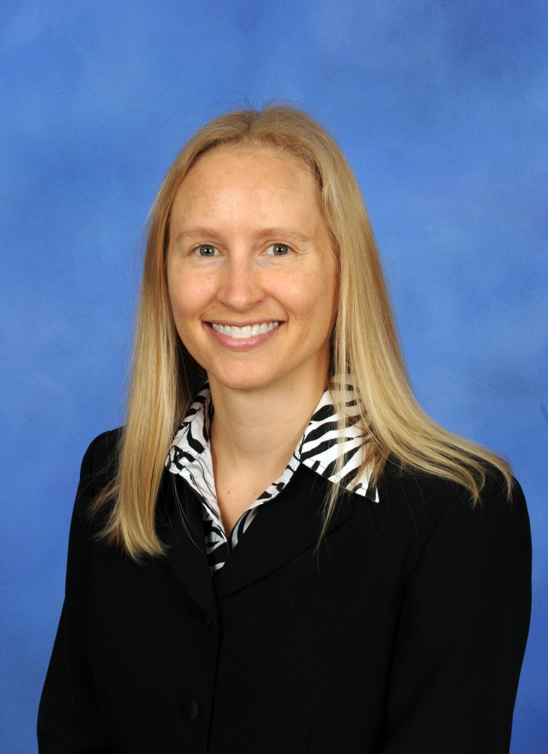 A blonde woman with a black jacket poses in for a headshot in front of a blue background.