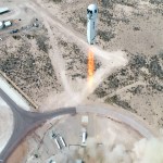 Aerial view of a desert area. Fire emerges from bottom of rocket rising from launchpad. Smoke and dust billow skyward.