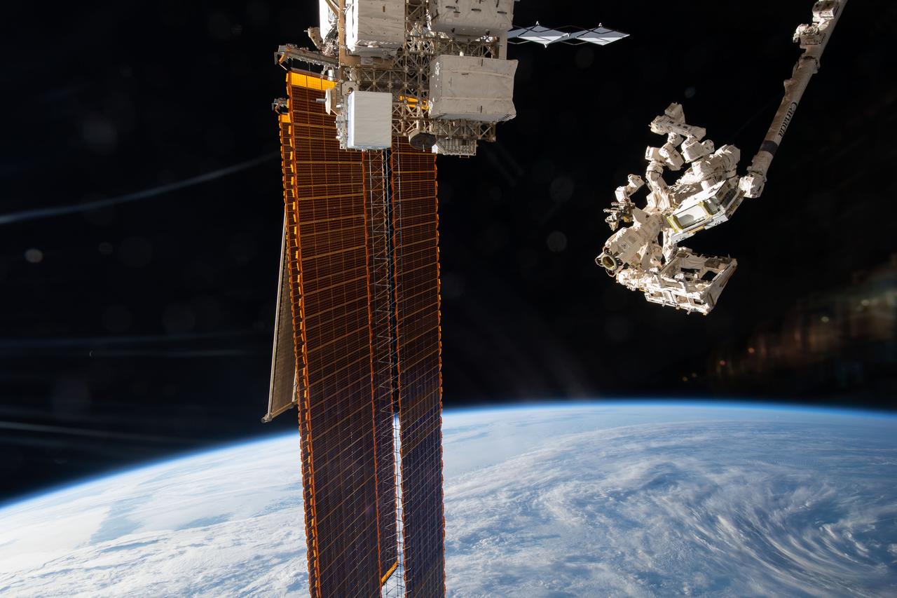 At right, the Canadarm2 robotic arm maneuvers the Earth Surface Mineral Dust Source Investigation, or EMIT, after retrieving it from the trunk of the SpaceX Dragon. The station's main solar arrays are seen extending from the port truss segment in the center of the photo. Earth is in the lower background while the blackness of space covers the upper background.