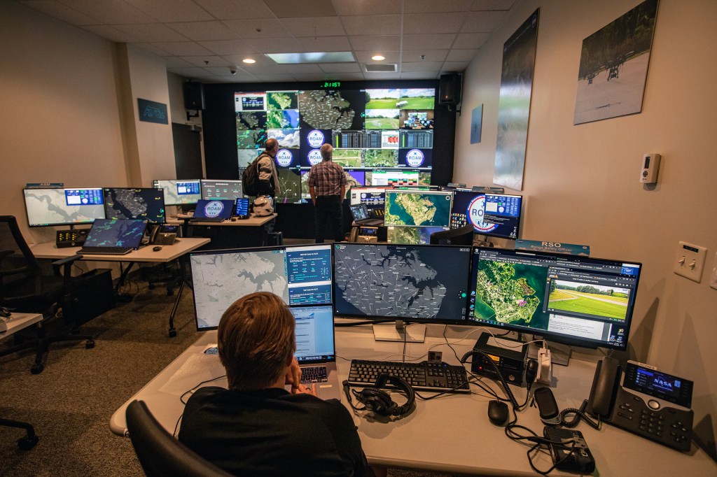 A room full of computer screens on tables and a far wall are watched by researchers monitoring the flight of small drones.