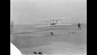 In this black and white photo, a white airplane with two sets of stacked wings with wires connecting them flies low to the ground. A man, Wilbur Wright, stands on the right with his back to the camera.