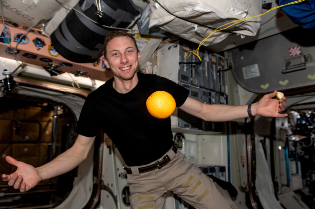 iss068e075550 (March 16, 2021) --- NASA astronaut and Expedition 68 Flight Engineer Woody Hoburg shows off a fresh orange, recently delivered aboard the SpaceX Dragon resupply ship, flying in microgravity aboard the International Space Station.