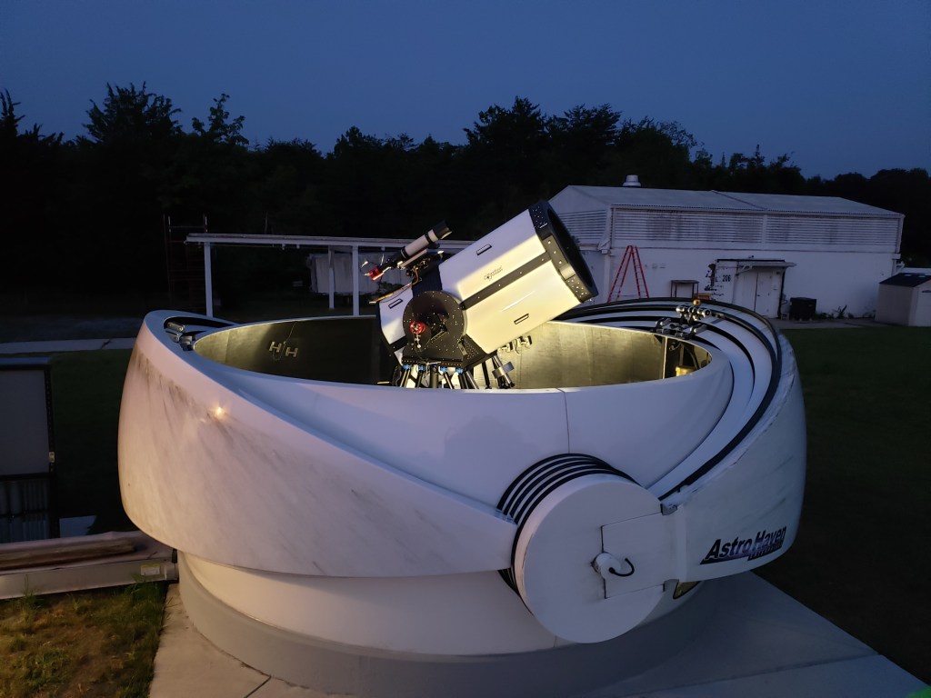 The Low-Cost Optical Terminal (LCOT), a white and black object resembling a telescope sits within a white shell with multiple sealable layers folded down to the sides. white buildings and dark trees line the background in this night scene.