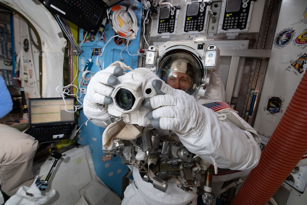 Inside the Quest airlock, NASA astronaut Nick Hague is fitted in a U.S. spacesuit and checks out a spacewalk camera days before beginning his first spacewalk.