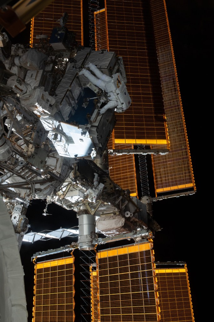 NASA astronauts Nick Hague (top) and Anne McClain (lower center) are dwarfed by the International Space Station's solar arrays as they work to swap batteries and install adapter plates during a six-hour, 39-minute spacewalk to upgrade the orbital complex's power storage capacity.