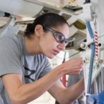 female astronaut in gray t-shirt and safety goggles working on a piece of hardware in a lab setting