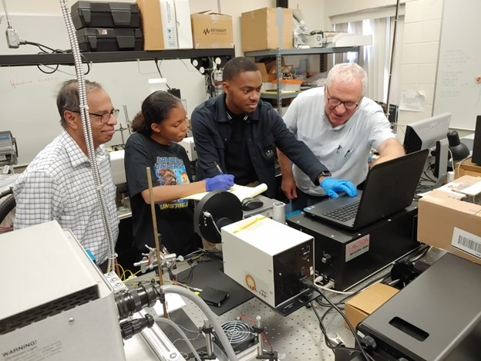Four people stand are gathered around a computer screen. The person on the left is Dr. Darayas Patel of Oakwood University; on the right is the President and CEO of SSSOT, Dr. Sergey Sarkisov. Two students stand between them, recording data from the computer, while Dr. Patel and Dr. Sarkisov guide them.