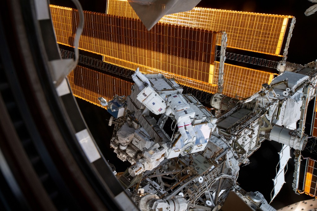 NASA astronauts Nick Hague (suit with no stripes) and Anne McClain (suit with red stripes) work to retrieve batteries and adapter plates from an external pallet during a spacewalk to upgrade the International Space Station's power storage capacity.