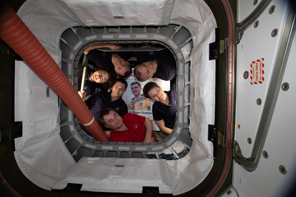 The six-member Expedition 59 crew poses for a portrait inside Northrop Grumman's Cygnus commercial space freighter dubbed the S.S. Roger B. Chaffee. Pictured in the center is a portrait of Chaffee who was a pilot for the Apollo 1 crew who lost their lives in a fire during a launch pad test of the Command Module at Kennedy Space Center. Clockwise from bottom are cosmonauts Alexey Ovchinin and Oleg Kononenko; NASA astronauts Christina Koch and Nick Hague; Canadian Space Agency astronaut David Saint-Jacques and NASA astronaut Anne McClain.