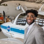 This is a photo of Brandon Sells, an aerospace engineer with the Aeronautics Systems Analysis Branch (ASAB) of the Systems Analysis and Concepts Directorate (SACD) at NASA Langley Research Center. Brandon is standing in front of a small white and blue plane with a propellor. A larger white plane can be seen in the background of the photo.