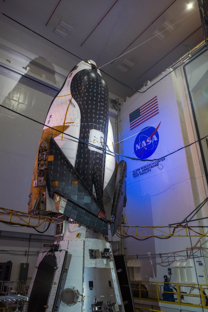 Nose-up and bathed in soft blue lights, Sierra Space’s Dream Chaser spaceplane and its Shooting Star cargo module cast dramatic shadows onto the walls of NASA’s Neil Armstrong Test Facility in Sandusky, Ohio. The spaceplane is black, white, and dark orange, and its wings are folded up. A NASA meatball and an American flag can be seen on the wall behind the spaceplane.