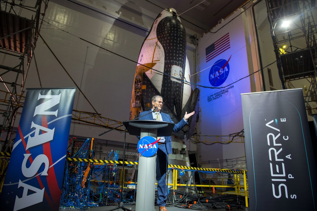 A man in a suit addresses a crowd of media inside the Mechanical Vibration Facility at NASA’s Neil Armstrong Test Facility. He stands at a podium that has a NASA meatball on it. On either side of him are banners that say “NASA” and “Sierra Space.” Behind him is the Dream Chaser spaceplane and its Shooting Star cargo module, nose-up and bathed in soft blue lights. Around the spacecraft are scaffolding and wiring, and a NASA meatball and an American flag can be seen on the wall.