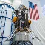 The Intuitive Machines Nova-C lander for the company's first Commercial Lunar Payload Services delivery is positioned before being encapsulated inside its launch fairing. The lander is yellow, grey, black, and white and an American flag and NASA and Intuitive Machines logos can be seen on the spacecraft’s surface. The background of the facility that the spacecraft is in is white, and a large American flag can be seen on the wall behind it.