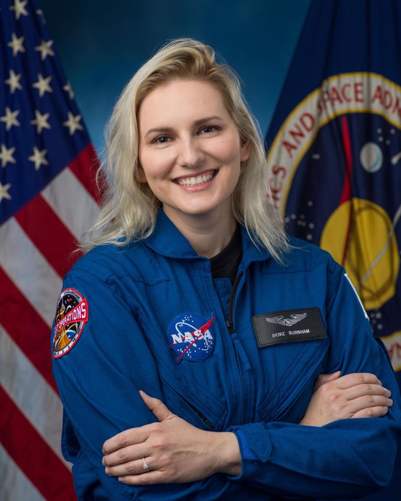 Astronaut Candidate Deniz Burnham smiles posing for an individual portrait. She wears a blue flight suit with her arms crossed standing infront the U.S. flag and NASA seal. Image Credit: NASA/Robert Markowitz