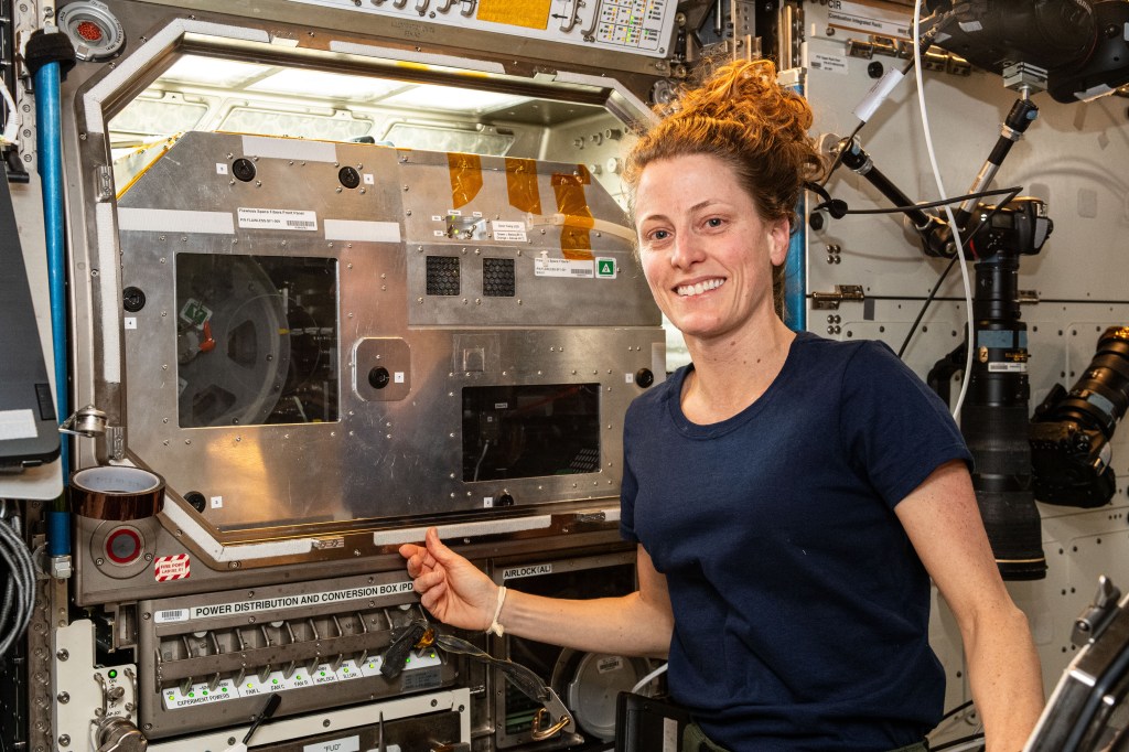 O’Hara, wearing a blue t-shirt with her hair in a ponytail, smiles at the camera and with her right hand reaches toward the Flawless Fibers hardware. Its large silver front panel has two windows, with a reel visible through the one on the left and controls through the one on the right.