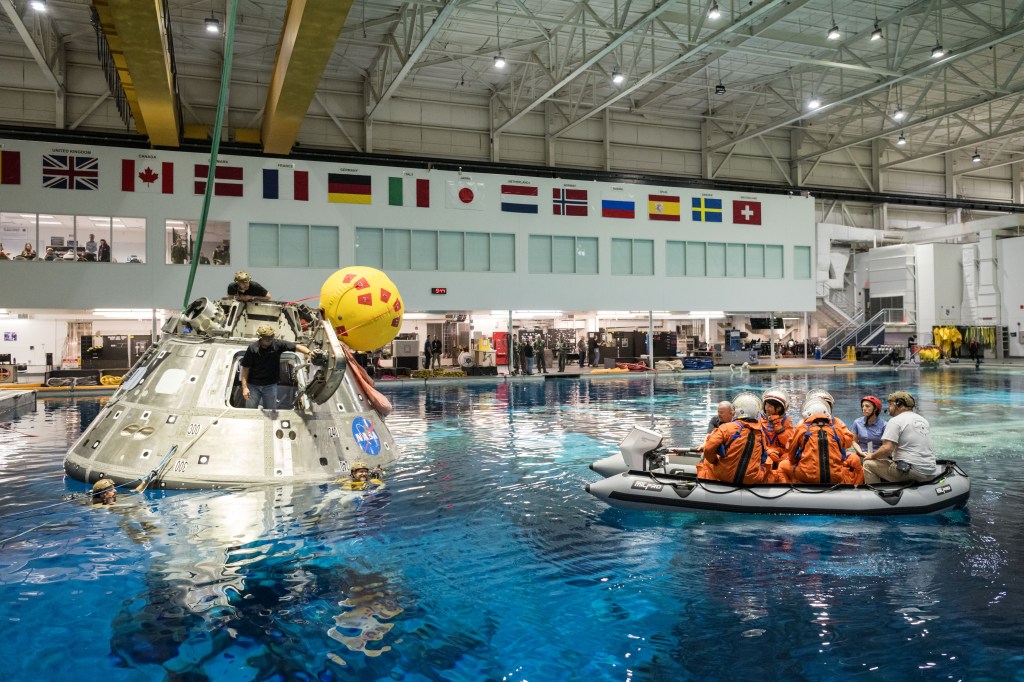 The Artemis II crew during their Orion Water Survival Demo in the Neutral Buoyancy Laboratory. While they are seated on an inflatable, they look towards the Orion Mockup that is floating inside the indoor pool. Credit: NASA/Josh Valcarcel