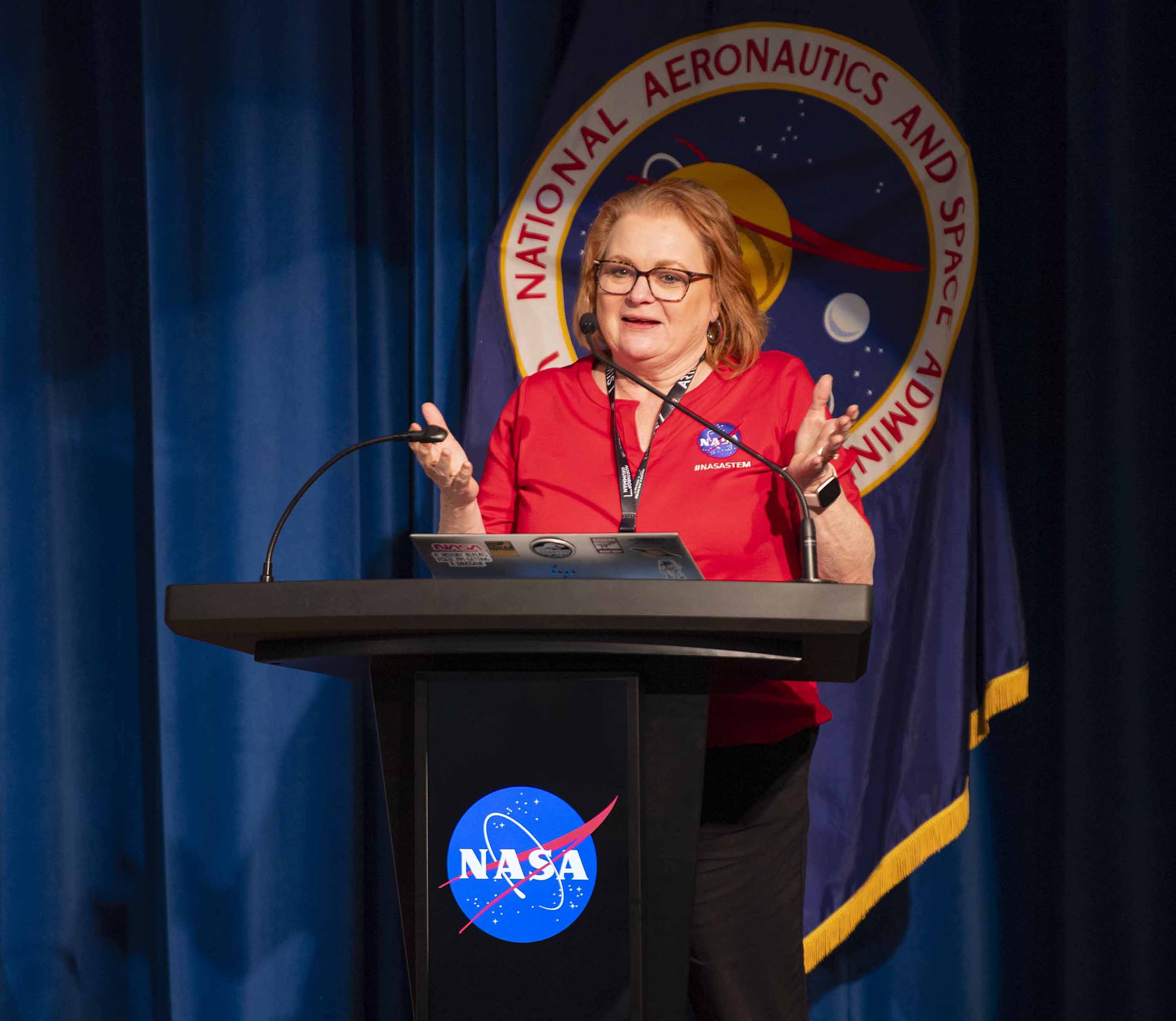 A woman, Next Gen STEM Project Manager Dr. Carrie Olsen, wearing a red polo with NASA meatball, speaks from behind the lectern