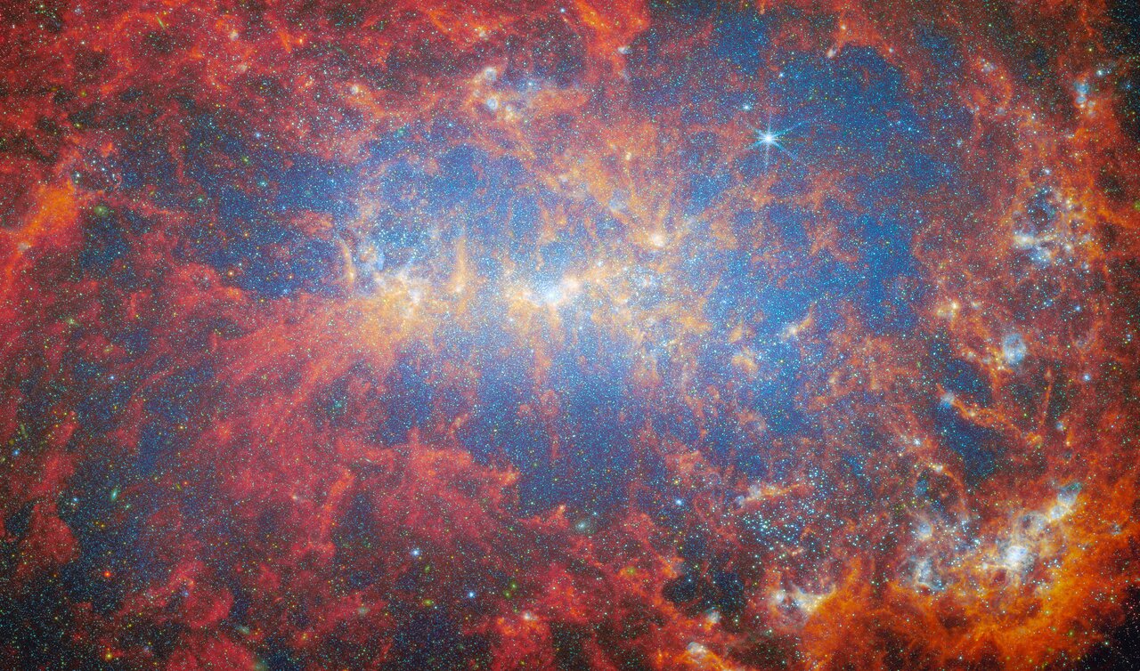 A close view of the central area of a dwarf galaxy. A huge number of stars fill the whole galaxy as tiny glowing points. They are brightest around the galaxy’s shining core. Thick clouds of gas and dust billow out across the scene, curling like moving flames. They glow in warm colours following their location: orange around the galaxy’s core, and around glowing star clusters in the bottom-left, and dark red elsewhere.
