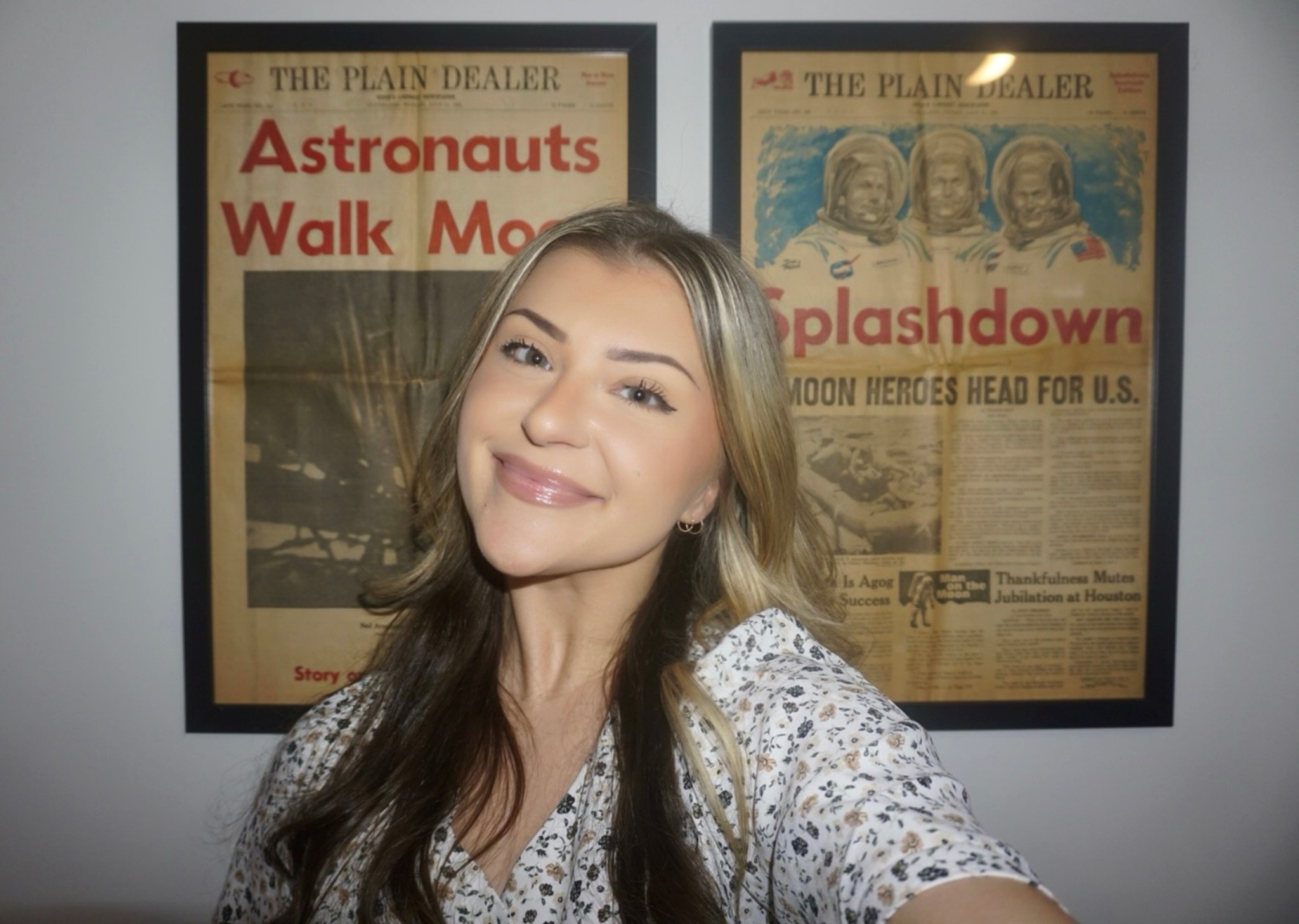 Cassi Meyer, wearing a white blouse with small flowers, poses in her selfie with two framed newspaper front pages hanging on the wall behind her.