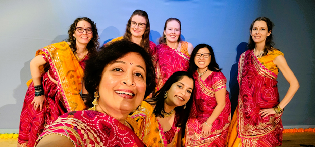 A group of seven diverse women wearing orange and pink saris prepare to perform a traditional dance during a Diwali celebration.
