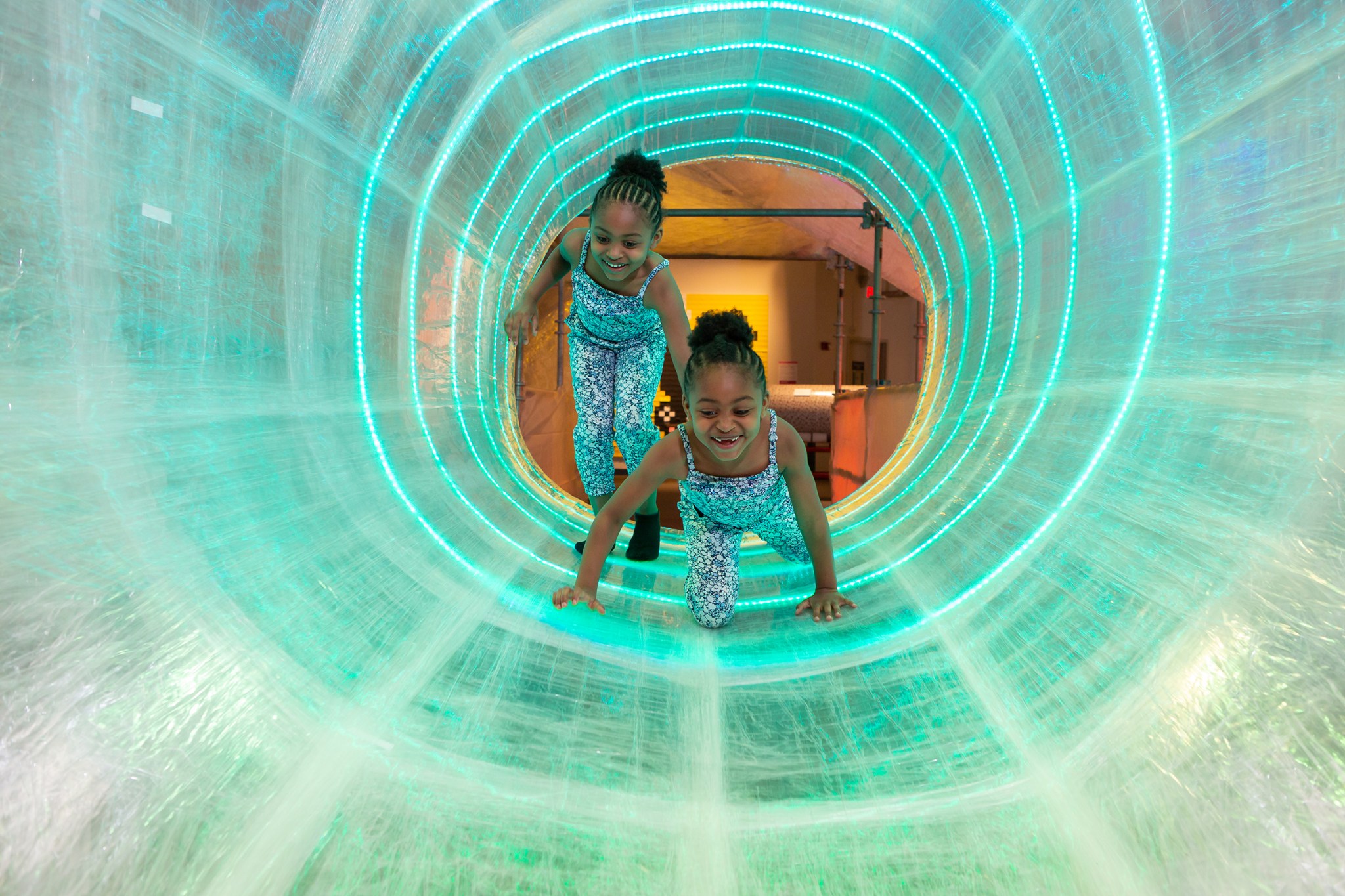 Two twin girls smile and laugh as they crawl through a large tunnel made of packing tape.