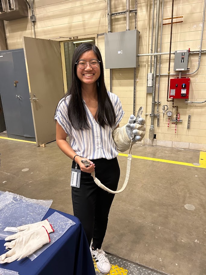 A young Asian American woman tries on a spacesuit glove while holding a tether used by astronauts during spacewalks.