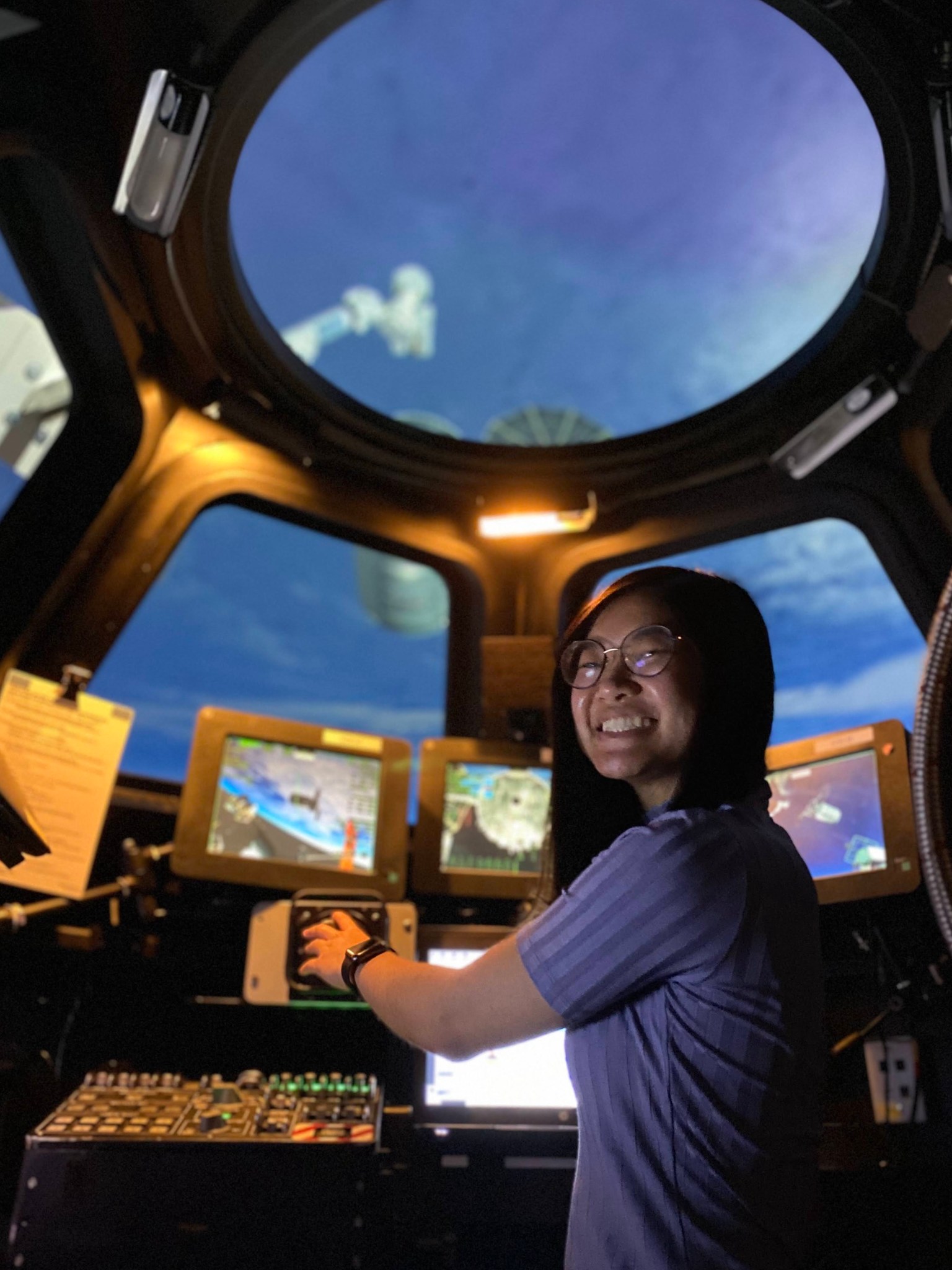 A young Asian American woman stands at the controls during a simulation inside a mockup of the International Space Station's cupola.