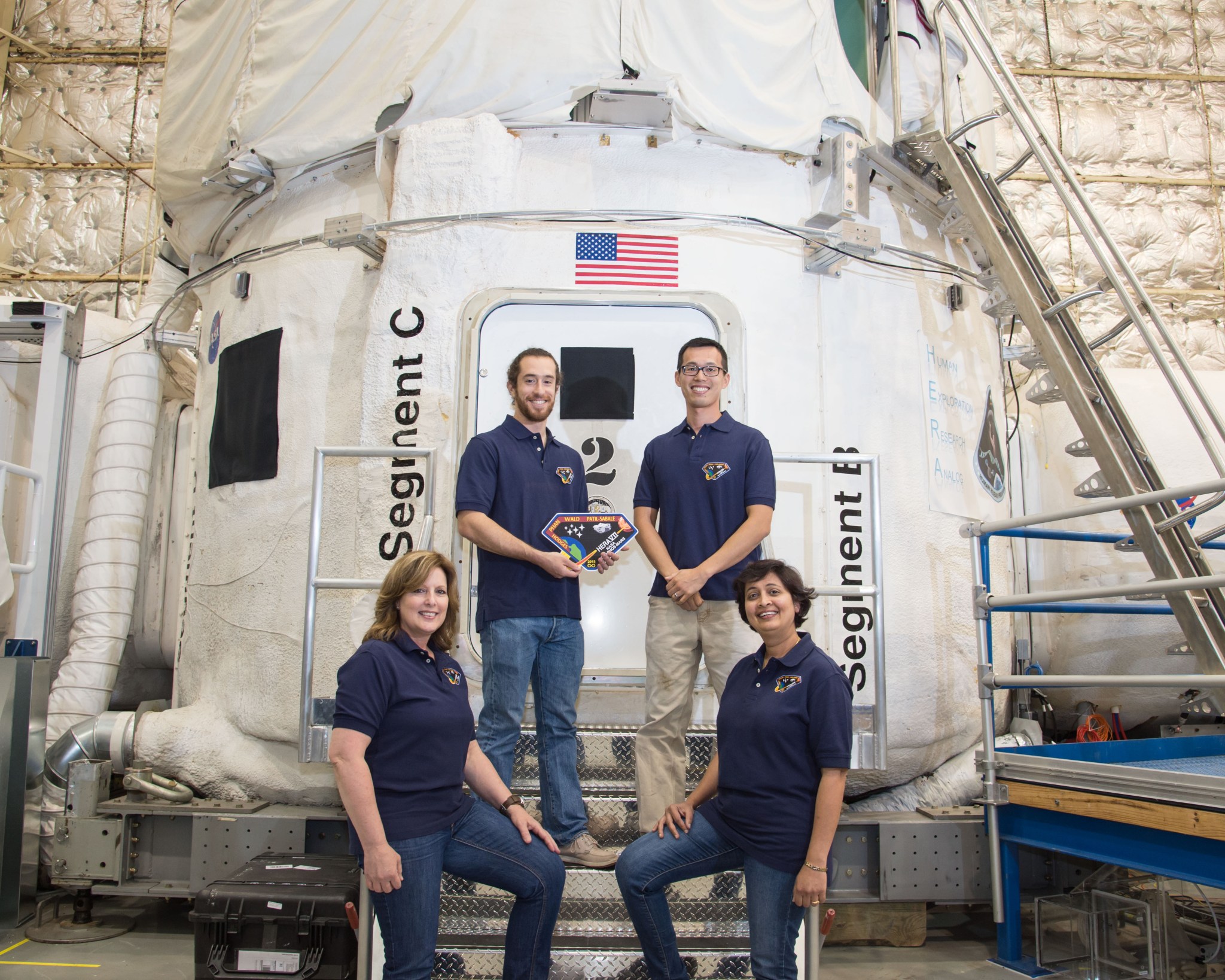 Two men and two women wearing matching blue polo shirts stand on the steps of a habitat designed to simulate a spacecraft flying to Mars.