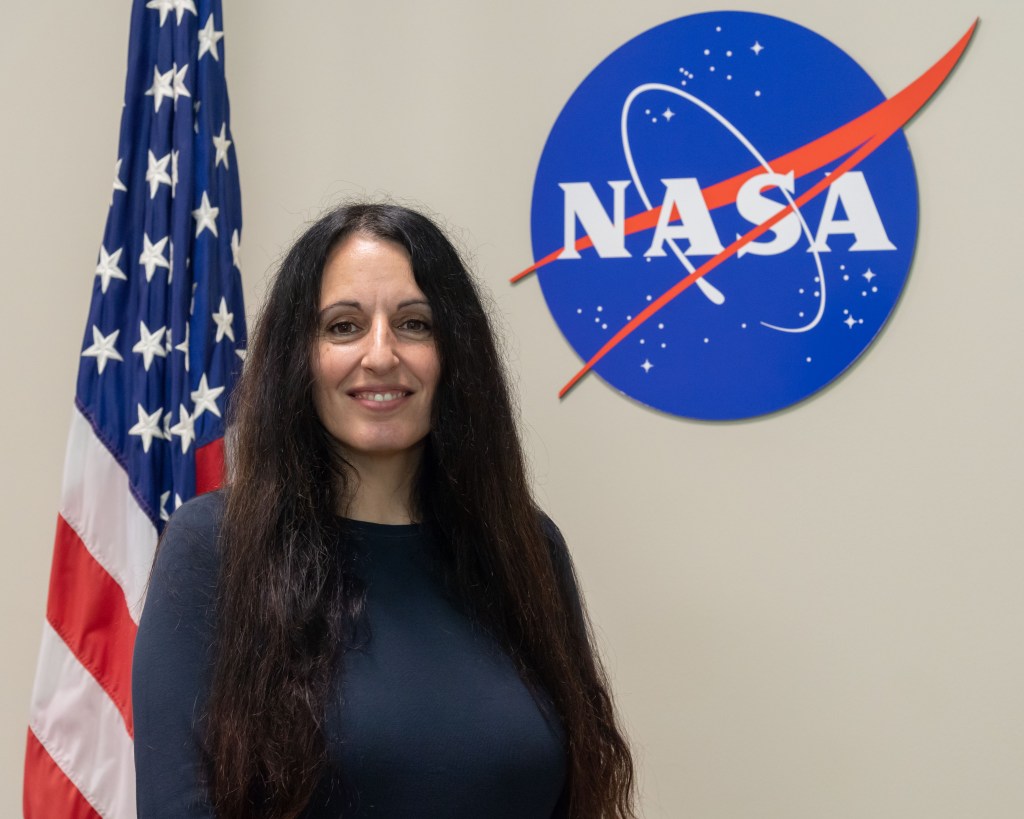 A woman with long dark hair, wearing a dark top, stands in front of the U.S. flag and a NASA logo on a beige wall.
