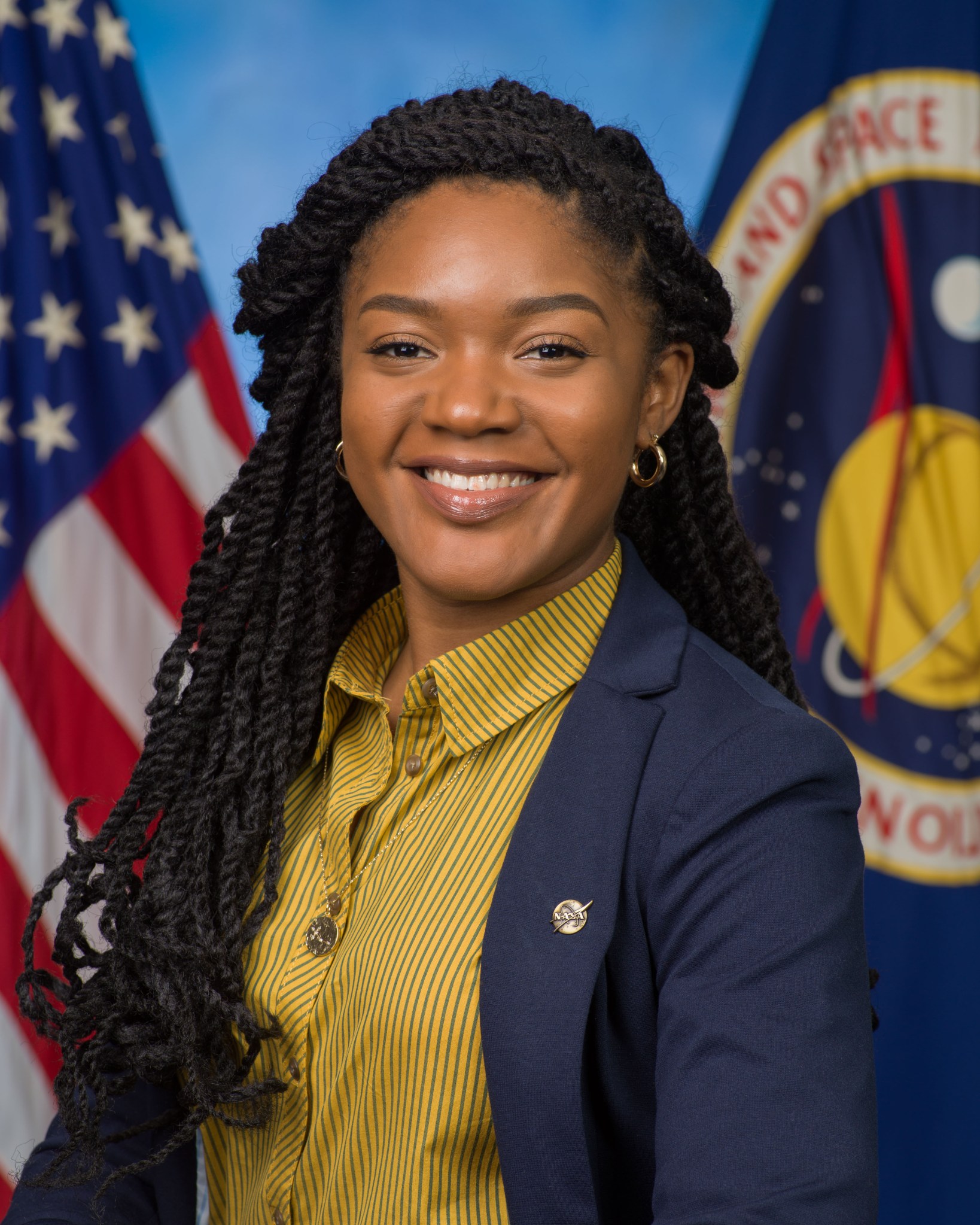 A woman wearing a yellow blouse and black blazer smiles in front of a blue background with two flags behind her, a U.S. flag on the left and a NASA flag on the right.