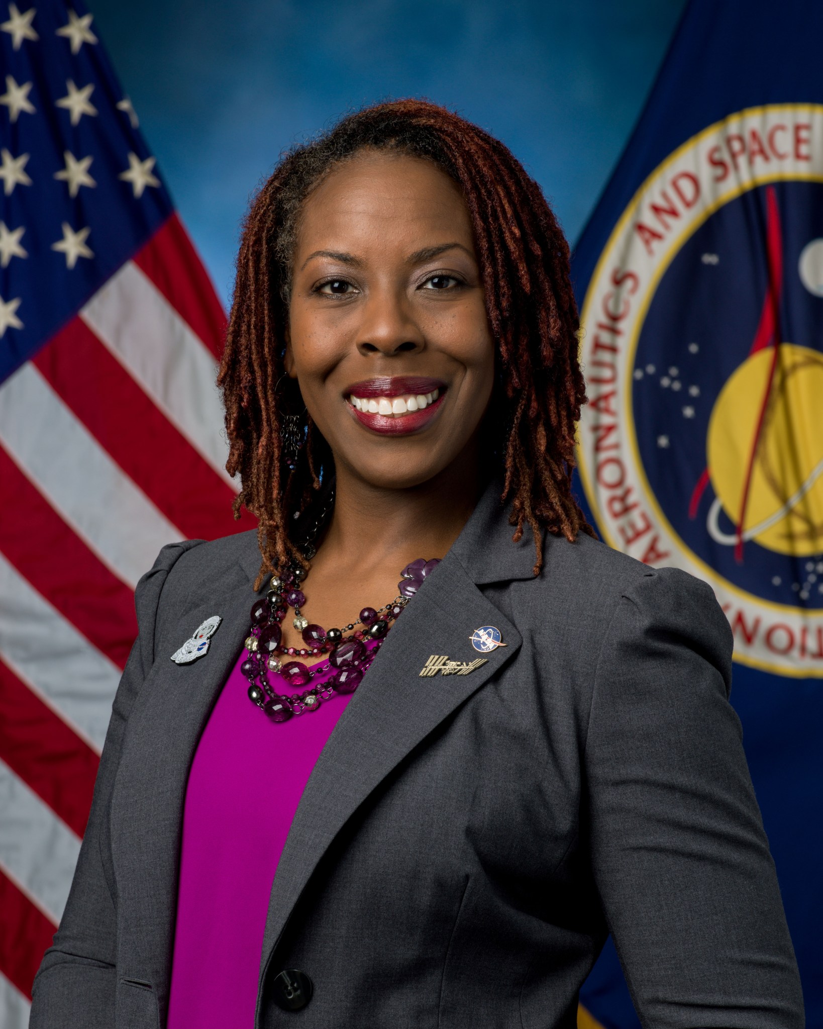 A woman wearing a grey blazer and a pink shirt smiles in front of a blue background with two flags behind her, a U.S. flag on the left and a NASA flag on the right.