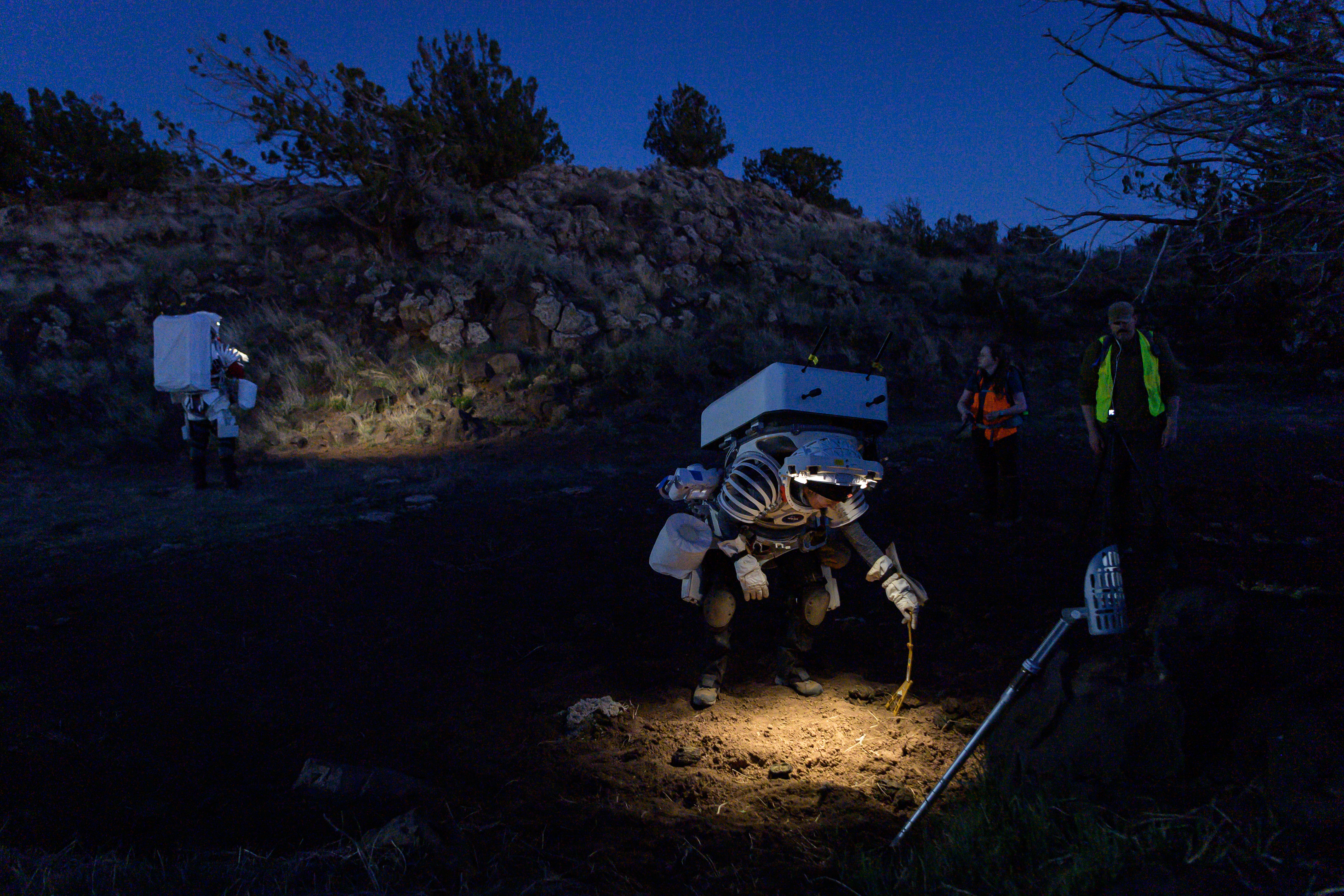 At night, an astronaut wearing white gear places a sample marker in the soil. The soil in front of her is lit up by the lights on the front of the open helmet. Two people in bright vests look on. In the background on the left, another astronaut wearing a similar outfit looks at another patch of soil.