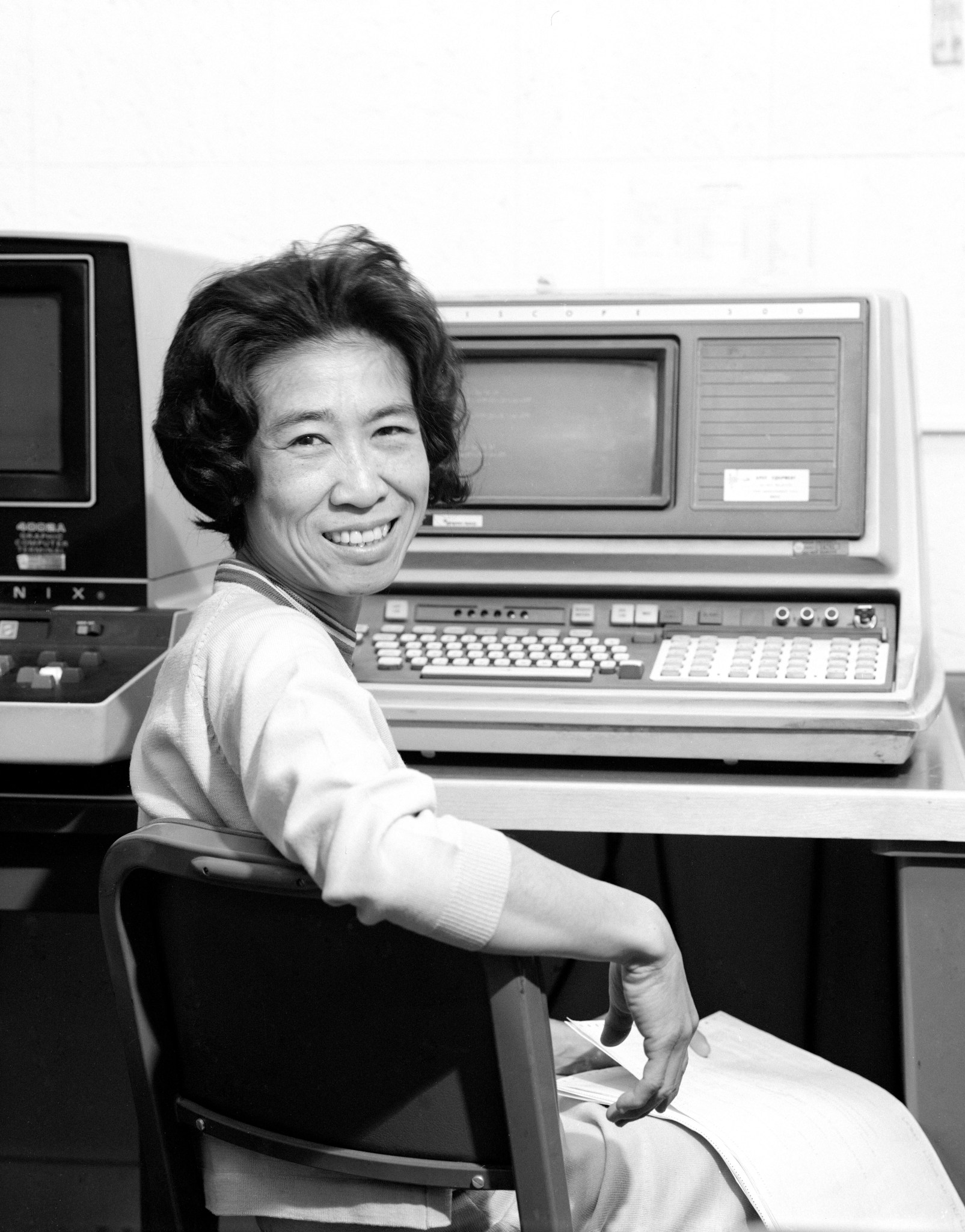 Helen Ling, an Asian woman, looks over her shoulder at the camera. Her arm is resting on the back of the chair she's sitting on. In front of her are several large computers.