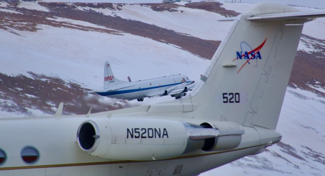 两架飞机在一座被雪和岩石覆盖的山前的图像。在前景中是一个白色喷流的尾端，填充了底部和右侧。NASA标志和编号520位于尾部。在这架喷气式飞机的后面，在图像的中间，另一架白色飞机起飞了。它是白色的，有一条蓝色的水平条纹，尾部有NASA的“蠕虫”标志。棕色和白色的山坡填满了画面的其余部分。