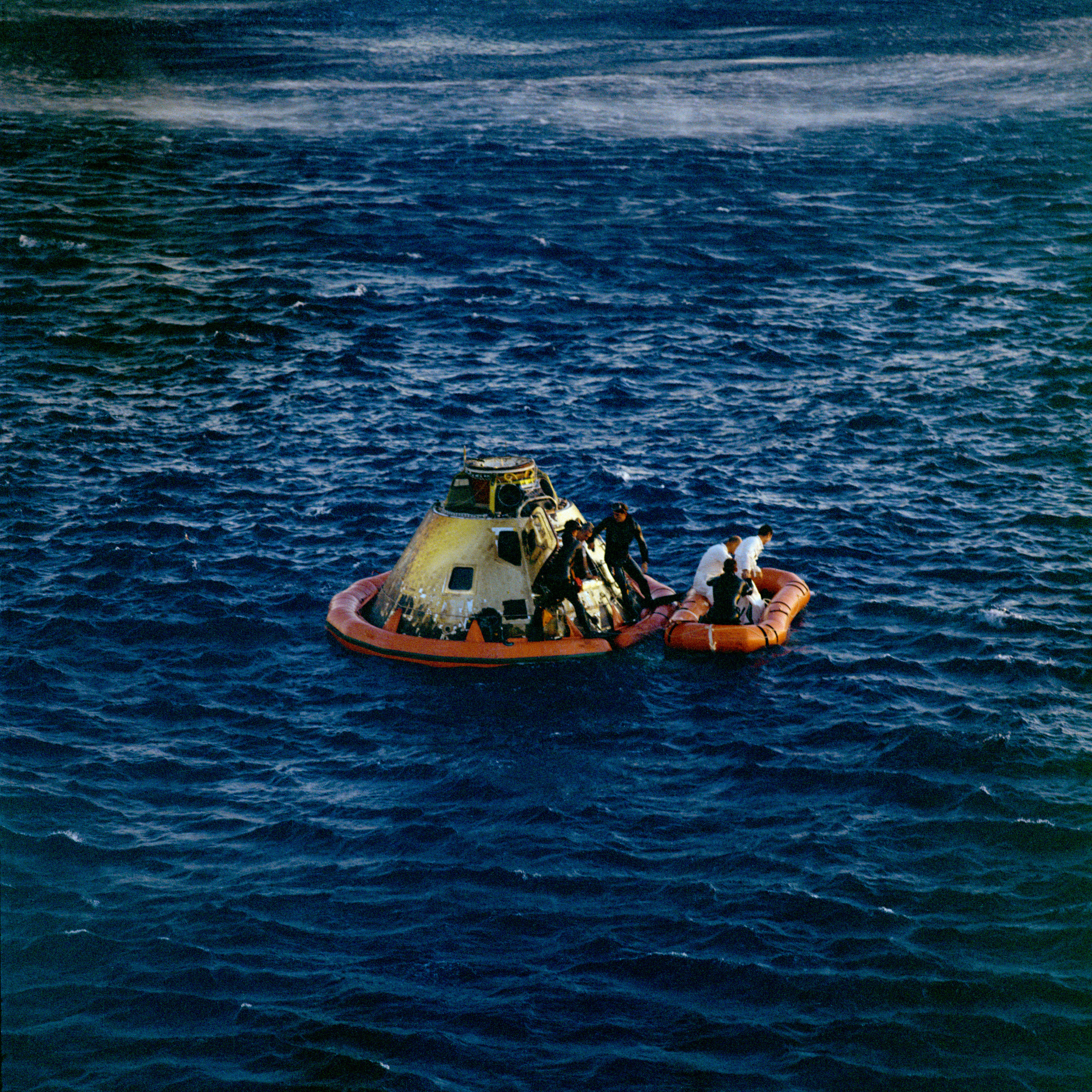 In the middle of a deep blue ocean, choppy with small waves, are two rafts. On the left is a larger orange raft that surrounds the Apollo 10 spacecraft. The spacecraft is a rounded pyramid shape and is painted white. Two men in black wetsuits look on as astronaut Eugene A. Cernan emerges from the spacecraft. Two other astronauts dressed in white are already on the smaller orange raft on the right, along with another person in a black wetsuit.