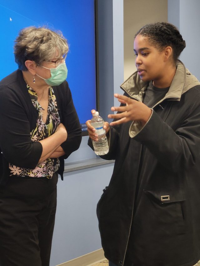 A woman wearing a mask stands listening to antother woman who is gesturing with her hands while holding a bottle of water.