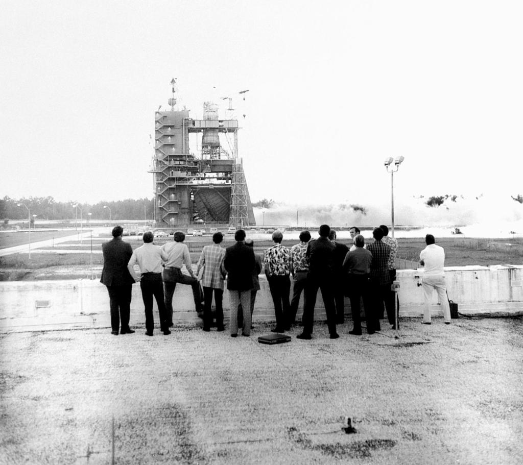 black and white photo captures the first static test-firing of the space shuttle main engine; a small crowd is seen observing the test from afar