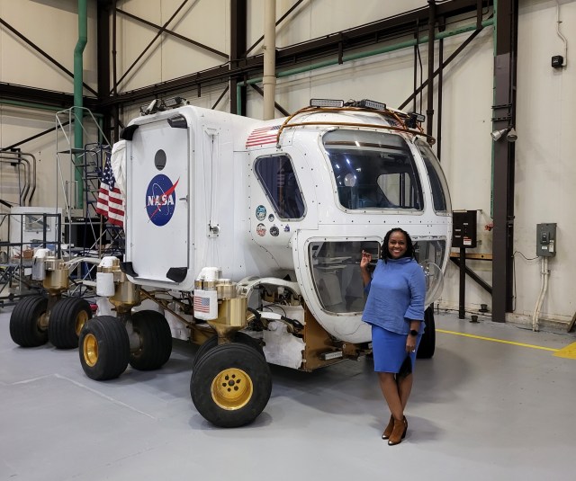 A woman stands smiling next to a NASA rover vehicle inside a large industrial facility. The woman is wearing a blue dress and brown shoes, and she is holding a small item in her hand. The American flag is visible in the background.